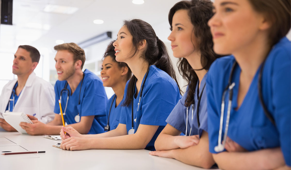 nurse-staffing A confident healthcare professional in blue scrubs standing with arms crossed, with a team of doctors and nurses in the background—representing the dedicated medical staffing services of Mid-South Care Services in Memphis, TN.